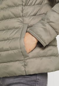Puffjacka i olivgrön, med quiltad struktur, som har en sidoficka och elastiska ärmslut. Materialet ser slätt och glänsande ut.