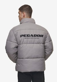 Grå pufferjakke med højt krave, der har sort "PEGADOR" tekst og "CERTIFICERET AUTENTISK STREETWEAR" på ryggen. Skinnende, kviltet tekstur.