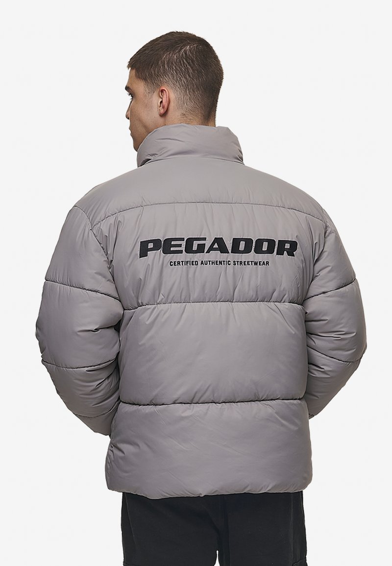 Pilkos pūkinės striukės su aukštu apykakle, ant kurios nugaros yra juodas užrašas "PEGADOR" ir "CERTIFIED AUTHENTIC STREETWEAR". Blizgi, siūta tekstūra.