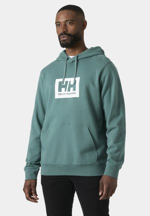 Hombre con sudadera con capucha Helly Hansen color verde azulado y bolsillo delantero, de pie contra un fondo liso gris claro, mirando ligeramente hacia la derecha.