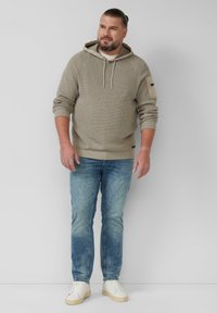 Beżowy dzianinowy hoodie z regulowanym ściągaczem, w połączeniu z jasnoniebieskimi dżinsami i białymi trampkami. Zauważalny detal w postaci naszywki na rękawie.