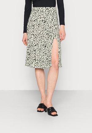 Femme portant une jupe midi blanche avec des taches abstraites noires et une fente sur la cuisse, associée à des sandales à talons noires et un haut noir.