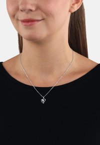 Silverhalsband i form av ett hjärta, tillverkat av metall med intrikata detaljer, uppvisas på en modellerad halslinje mot en svart skjorta.