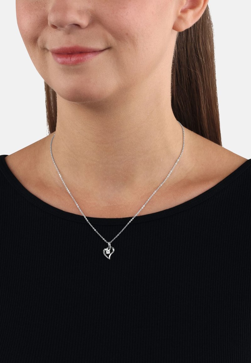 Silverhalsband i form av ett hjärta, tillverkat av metall med intrikata detaljer, uppvisas på en modellerad halslinje mot en svart skjorta.