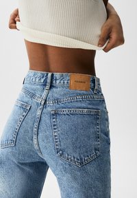Persona levantando una blusa ribeteada crema, usando jeans de mezclilla azul de cintura alta con una etiqueta marrón de Pull&Bear en la parte trasera de la cintura.