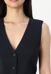 Black sleeveless garment with deep V-neckline, featuring three black buttons down the center, made of a smooth, textured fabric.