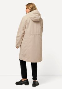 Helle beige Kapuzenjacke mit tailliertem Schnitt und langen Ärmeln. Das Material wirkt glatt und wasserabweisend, ohne sichtbare Muster.