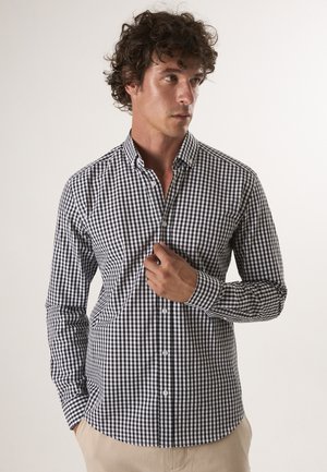 Mand med krøllet hår, der justerer kraven på en sort-hvid ternet skjorte med knapper, iført beige bukser mod en ensfarvet baggrund.