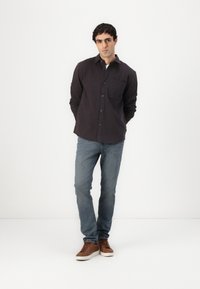 Camicia scura con colletto e taschino sul petto, abbinata a jeans azzurri e sneakers marroni, in piedi su uno sfondo bianco.