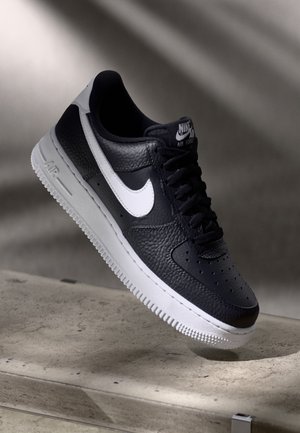 Černobílá teniska Nike Air Force 1 s bílou podrážkou, vznášející se nad betonovým povrchem ve měkkém osvětlení.