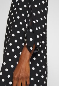 Blusa negra con lunares blancos, de corte holgado, con mangas atadas y una textura de tela suave. Enfócate en el diseño de las mangas y el contraste del patrón.