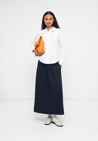 Camicia bianca a maniche lunghe con bottoni abbinata a una lunga gonna plissettata blu scuro. Tiene in mano una borsa a tracolla triangolare arancione. Scarpe di colore chiaro.