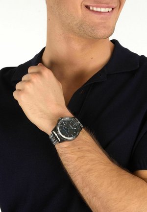 Mann, der ein schwarzes Poloshirt trägt, lächelt und zeigt eine silberne Metallarmbanduhr mit schwarzem Zifferblatt und blauen Akzenten an seinem linken Handgelenk.