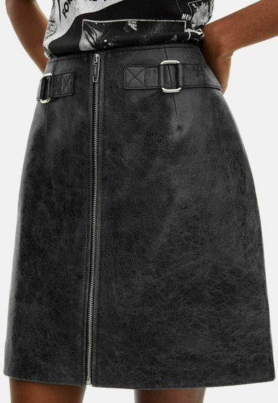 Zwarte faux leren rok met een vooraan aangebrachte rits, twee zijgespen en een textuur. Heeft een gestructureerde A-lijn vorm en minimale accenten.