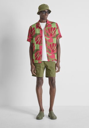 Grand homme portant un chapeau bob vert, des lunettes de soleil noires, une chemise rouge et verte à motifs par-dessus un tee-shirt blanc, un short vert olive et des chaussures slip-on olive.