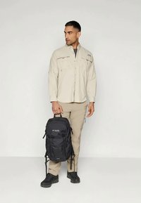 Black backpack with zippers and logo, worn over a light beige long-sleeve shirt and tan cargo pants, paired with black hiking shoes.