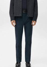 Mann, der eine dunkelgraue Jacke, einen schwarzen Gürtel, marineblaue karierte Hose und schwarze Lederschuhe trägt und vor einem schlichten Hintergrund steht.