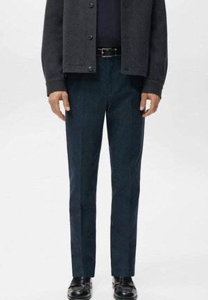 Man wearing dark grey jacket, black belt, navy blue checked pants, and black leather shoes, standing against a plain background.