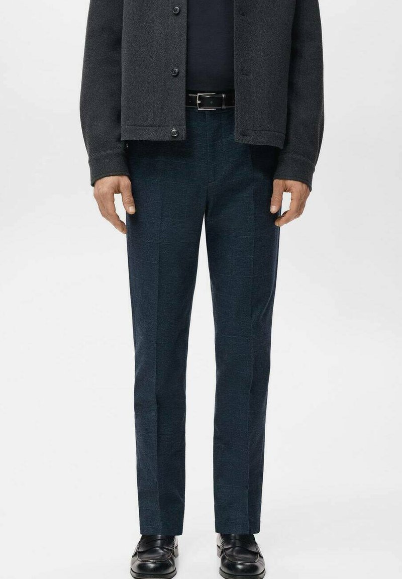 Mann, der eine dunkelgraue Jacke, einen schwarzen Gürtel, marineblaue karierte Hose und schwarze Lederschuhe trägt und vor einem schlichten Hintergrund steht.