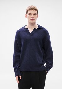 Πλεκτό πουλόβερ σε ναυτικό μπλε με V λαιμόκοψη και μεγάλο γιακά, με σχεδιασμό χαμηλωμένων ώμων και ριμπ μανσέτες και τελείωμα. Φοριέται πάνω από μαύρο παντελόνι.