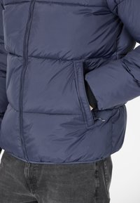 Veste matelassée bleu marine avec sections matelassées, fermeture éclair complète, poches latérales et une finition lisse. Conçue pour la chaleur et le confort.