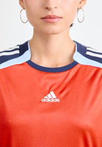 Raudona sportinė marškinė su mėlynais ir tamsiai mėlynais akcentais, su balta Adidas logotipo emblema ant krūtinės ir lygiu, blizgiu audiniu.