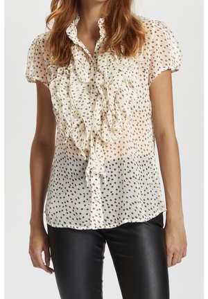 Blouse en crêpe, couleur crème, avec un devant à volants, ornée de pois marron, manches courtes bouffantes et fermeture à boutons. Portée avec un pantalon en cuir noir.