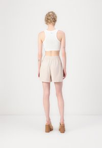 Un crop top blanc sans manches, un short beige texturé et des sandales à talons épais couleur beige. Modèle vu de dos sur un fond uni.
