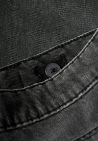 Tmavě šedá džínová látka s hladkou texturou, obsahující černé tlačítko se čtyřděrovým designem a viditelným prošitím podél okraje.