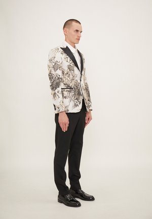 Blazer bianco con motivo floreale nero, colletto in satin nero, abbinato a pantaloni neri e scarpe nere lucide. Il modello è in posizione profilo.