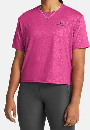 Osoba v jasně růžovém crop tričku Under Armour s jemnými vzory a šedých elastických legínách, stojící s uvolněnými pažemi.