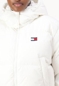 Hvit pufferjakke med høy krage, glidelås foran og brodert "TOMMY JEANS"-logo med røde og marineblå striper på brystet.