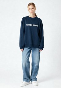 Sweat-shirt bleu marine avec le texte blanc "FASHION CRIMES". Comprend des poignets et un ourlet côtelés, associé à un jean bleu clair à coupe ample et des baskets blanches.