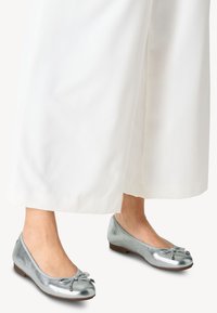 Ballet flats in argento metallizzato con punta arrotondata, tacco basso e un fiocco sul davanti. Indossati con pantaloni bianchi fluidi.