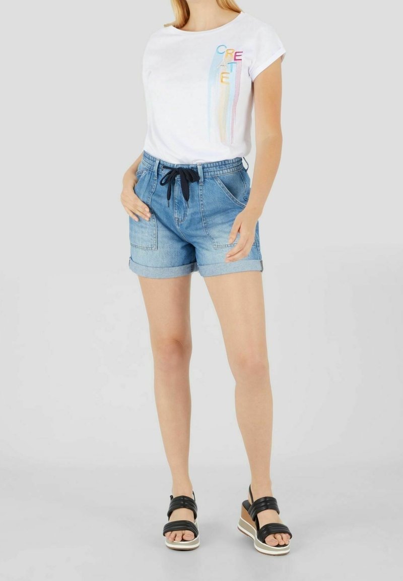 Valkoinen t-paita, jossa on "CREATE" -grafiikka, yhdistettynä vaaleansinisiin farkkushortseihin, joissa on kiristysnauha, käännetyt hihat ja sivutaskut. Mustat alustan sandaalit.