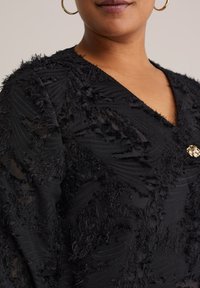Top in tessuto nero textured con design a strati, dettagli frangiati e un bottone decorativo dorato all'occorrente.