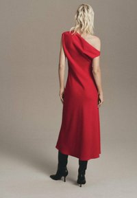 Vestido rojo de un solo hombro con tela suave y drapeada. Silueta larga y fluida que llega a la mitad de la pantorrilla. Combinado con botas negras de tacón alto.