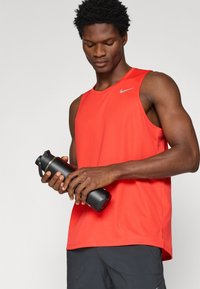 Piros ujjatlan sportos trikó, szürke logóval, mindkét kezében egy fekete vízpalackot tart. Sötét rövidnadrág és semleges háttér.