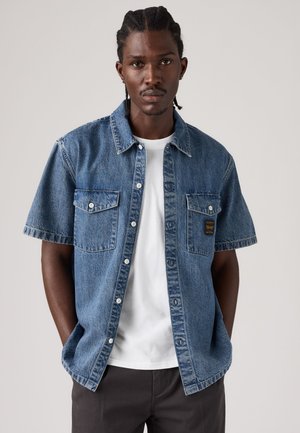 Mand med flettet hår iført en kortærmet denimskjorte over en hvid t-shirt, stående mod en ensfarvet lys baggrund.
