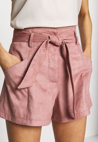 Růžové šortky z umělé suédé s páskem na zavázání, bočními kapsami a hladkým povrchem, mají vysoký pas a široký střih nohavic.