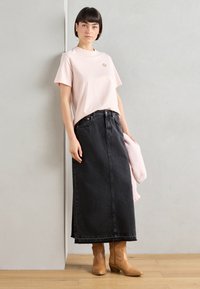 T-shirt in cotone rosa chiaro con una piccola grafica, abbinato a una lunga gonna di jeans nera. Stivaletti marroni con tacco completano l'outfit.