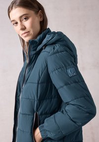 Giacca a vento teal con cuciture orizzontali, cappuccio e tasche con zip. Patch con logo sul braccio sinistro, texture liscia e stile aderente.