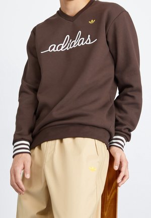 Henkilö pukeutuneena ruskeaan Adidas-collegepaitaan, jossa valkoinen käsinkirjoitettu logo, ja beigeihin housuihin, joissa pieni keltainen Adidas-logo, seisoo valkoista taustaa vasten.