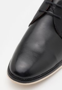 Scarpa in pelle nera con una superficie liscia e lucida, punta rotonda e suola beige chiaro. Presenta un pannello in rete nera testurizzata vicino ai lacci.