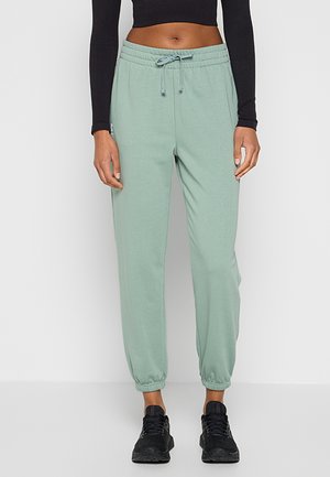 Donna con pantaloni della tuta verde chiaro con coulisse, top corto nero e sneakers nere, in piedi davanti a uno sfondo chiaro e uniforme.