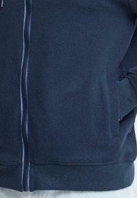 Sweat à capuche zippé bleu marine avec tissu texturé et surpiqûres bleues contrastantes. Comprend deux poches latérales et un ourlet côtelé.