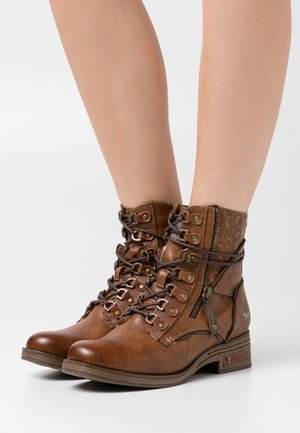Brown leather ankle boots with lace-up front, decorative embossed pattern, side zipper, and low stacked heel. Features metal eyelets and accents.