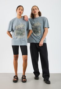 Camisetas gráficas grises con un diseño de serpiente, combinadas con pantalones cortos de bicicleta negros acanalados y sandalias negras en dos modelos. Fondo blanco.