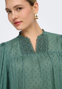 Γυναίκα που φοράει μπλούζα πράσινου χρώματος με υφή και λεπτομέρειες δαντέλας στο γιακά, καθώς και χρυσούς κρίκους στα αυτιά, κοιτάζοντας ελαφρώς προς το πλάι.