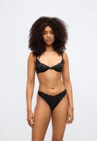 Súprava čiernej čipkovanej spodnej bielizne s balconette podprsenkou s kostrou a zodpovedajúcimi tangami. Zložitý kvetinový čipkovaný vzor a zvlnené okraje.